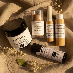 Op de afbeelding: Een collectie Odin Hollow huidverzorgingsproducten, waaronder een ronde pot met een zwarte deksel, een doorzichtige stick-verpakking en drie cilindrische tubes met witte etiketten en zwarte tekst op een beige textiel.