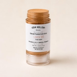 Könnte beinhalten: Ein zylindrischer Creme-Foundation-Stift in einem beigen Farbton mit einer transparenten Kunststoffbasis. Das weiße Etikett trägt die Aufschrift ODIN HOLLOW Beauty, CREAM FOUNDATION STICK, LINEN 100, FAIR IVORY, BOTANICAL OILS + MINERAL PIGMENTS, 17 g, ODIN HOLLOW BEAUTY, BELTON, SC USA.