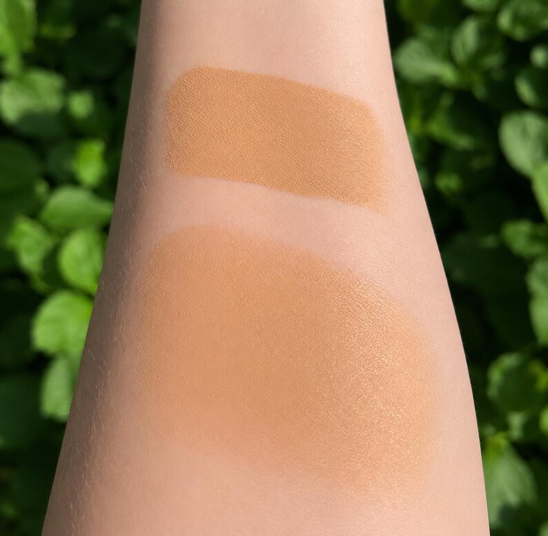 May include: Two tan foundation swatches on an arm, one lighter and one darker. The top swatch is a lighter shade of tan, and the bottom swatch is a darker shade. Green foliage is blurred in the background.