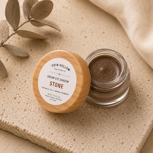Pode incluir: Um pequeno frasco de vidro com sombra cremosa na cor Stone, com tampa de madeira clara. O rótulo branco diz Odin Hollow Soap & Tallow Co., Cream Eye Shadow, Stone, Botanical Oils + Mineral Pigments, Net Wt. 0.17 oz | 5 g, Made in Small Batches in Texas.
