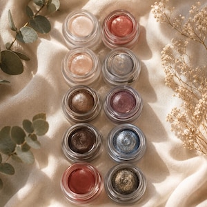 May include: Ten small, round, clear glass jars filled with cream-based eyeshadows in various metallic and shimmer finishes, including shades of copper, gold, bronze, plum, blue, and silver, arranged in two vertical columns on a soft fabric.