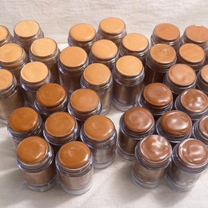 May include: A selection of small, clear jars containing different shades of foundation. The jars have brown lids, and the foundation colours vary from light beige to dark brown. The jars are arranged on a white surface.