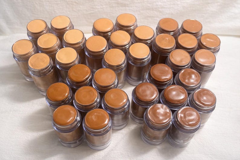May include: A selection of small, clear jars containing different shades of foundation. The jars have brown lids, and the foundation colours vary from light beige to dark brown. The jars are arranged on a white surface.