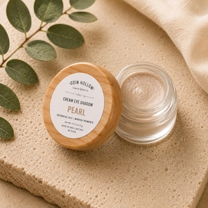 Puede incluir: Un pequeño frasco de vidrio con sombra de ojos en crema color perla y tapa de madera clara. La etiqueta dice ODIN HOLLOW Soap & Tallow Co. CREAM EYE SHADOW PEARL BOTANICAL OILS + MINERAL PIGMENTS Net Wt. 0.17 oz | 5 g.