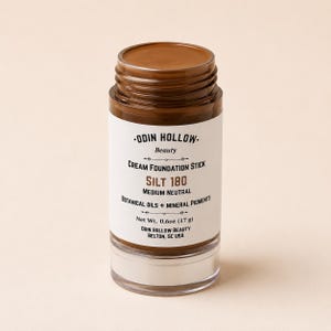May include: A cylindrical cream foundation stick in the shade Silt 180, medium neutral. The white label features black text reading Odin Hollow Beauty, Cream Foundation Stick, Silt 180, Botanical Oils + Mineral Pigments, and Net Wt. 0.6oz (17 g).