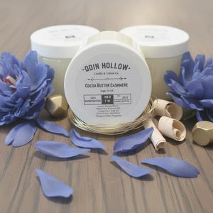 May include: Three white jars of sugar scrub with lids, labeled "Cocoa Butter Cashmere." The jars are surrounded by blue flower petals, blue flowers, gold geometric shapes, and wood shavings. The jars are 7 oz.
