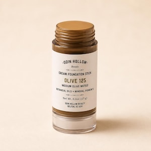 Puede incluir: Una barra de base de maquillaje en crema cilíndrica en el tono Olive 125. La etiqueta blanca presenta el texto Odin Hollow Beauty, Cream Foundation Stick, Olive 125, Medium Olive Muted, Botanical Oils + Mineral Pigments y Net Wt. 0.6oz (17 g).