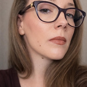 May include: Close-up of a person wearing dark purple eyeglasses with a gold accent on the temples. The person has light brown hair and is wearing brown lipstick. The background is a neutral colour.
