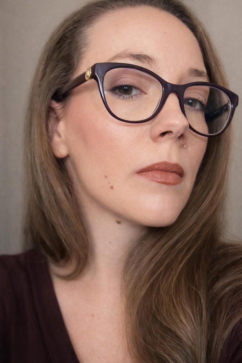 May include: Close-up of a person wearing dark purple eyeglasses with a gold accent on the temples. The person has light brown hair and is wearing brown lipstick. The background is a neutral colour.