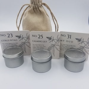 May include: Three silver metal candle tins with labels that read "No. 23 Citrus Petals", "No. 25 Calming Lily", and "No. 31 My Oud". The tins are sitting on a white surface next to a small, beige cloth bag.