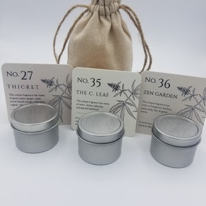 May include: Three small silver tins with labels that read "No. 27 Thicket", "No. 35 The C. Leaf", and "No. 36 Zen Garden". The tins are sitting on a white surface in front of a small, beige cloth bag.