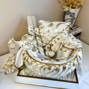 May include: A white and gold decorative basket filled with various items. The basket is adorned with gold coin-like embellishments and trim. Items include candles, a pillow, and decorative fabric. A vase with dried flowers is in the background.