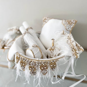 Pode incluir: Uma tigela de cerâmica branca com uma borda dourada e uma borda de penas brancas. A tigela está cheia de almofadas de cetim branco e uma boa de penas brancas. As almofadas e a boa são decoradas com strass dourados e transparentes.