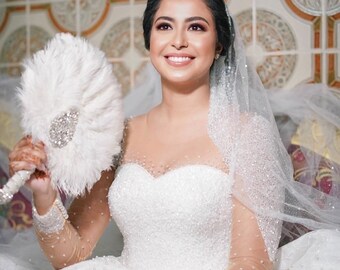 Könnte beinhalten: Eine Braut in einem weißen, perlenbesetzten Brautkleid mit langem Schleier und einem weißen Federfächer. Die Braut lächelt und blickt nach oben.