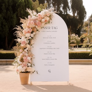 May include: A white archway sign with the text "Unser Tag" and the names "Celine & Sascha" followed by the date "24.06.2023". The sign lists the wedding day schedule with times and locations for the welcome drink, ceremony, appetizer and wedding cake, dinner, sweet buffet, and party.
