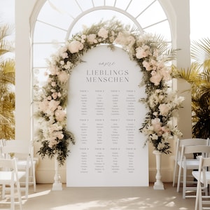 May include: A white seating chart with the text "unsere LIEBLINGS MENSCHEN" at the top. The chart lists names under different table numbers. The chart is decorated with a floral archway made of white and pink flowers.