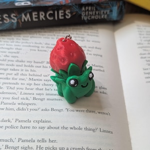 Puede incluir: Un llavero de criatura de fresa impreso en 3D. La parte superior de fresa roja se asienta sobre un cuerpo verde con grandes ojos blancos. El llavero está en una página de libro.
