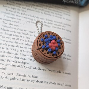 May include: A brown waffle pendant with blueberries and a red center, attached to a silver chain. The pendant is a miniature replica of a waffle, with detailed textures and colorful accents. The background is a book.