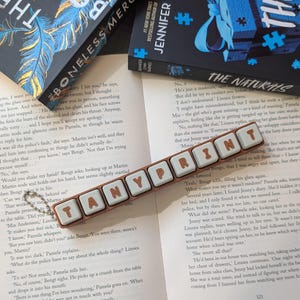 Puede incluir: Un llavero rectangular marrón con una fila de piezas cuadradas de color azul claro con las letras marrones TANYPRINT en relieve. El llavero incluye una pequeña cadena de bolas de metal plateado en un extremo, colocado sobre un libro abierto.