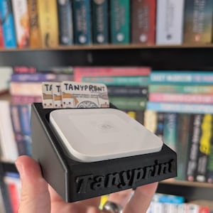 Può includere: Un supporto nero per lettore di carte stampato in 3D con la scritta "Tanyprint" sul davanti. Il supporto contiene un lettore di carte bianco e una carta con la scritta "T T TANYPRONT". Librerie sullo sfondo.