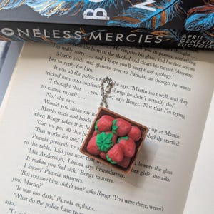 Puede incluir: Un dije de fresas en miniatura en una pequeña caja de madera, sujeto a una cadena plateada. Las fresas rojas tienen hojas verdes. El dije descansa sobre un libro abierto con el título "Boneless Mercies".