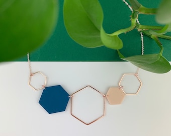 May include: A geometric necklace featuring a silver chain and a series of connected hexagonal shapes. The necklace includes a navy blue, a beige, and a clear hexagon, with gold-colored metal frames. The background is white and green.