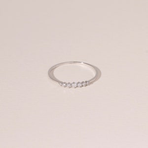 May include: A delicate silver ring featuring a row of small, sparkling gemstones. The ring has a thin band and the gemstones are evenly spaced across the top. The ring is displayed on a neutral background, highlighting its simple elegance.