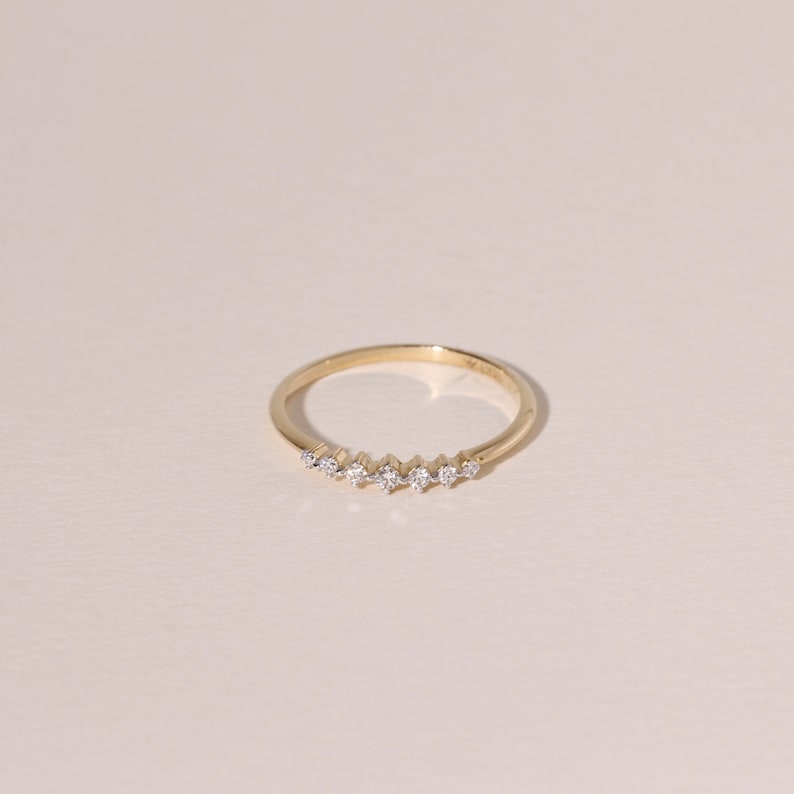 May include: A delicate gold ring featuring a series of small, sparkling gemstones set in a row. The ring has a thin band and a minimalist design, perfect for everyday wear. The gemstones are evenly spaced and catch the light, adding a touch of elegance.