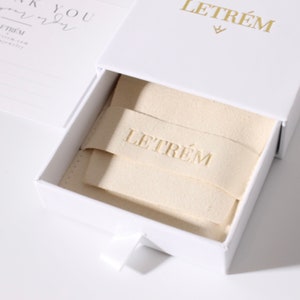 May include: Open white jewelry box with a pull-out drawer. The box has gold lettering that reads "LETR&Eacute;M" on the top and inside. A cream-colored fabric insert is inside the box, also with the name "LETR&Eacute;M" embossed on it. A white ribbon is attached to the drawer.