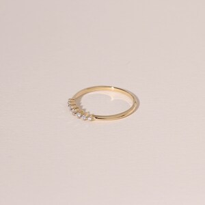 May include: A delicate gold ring with a row of small, clear gemstones set along the top. The ring has a thin band and a polished finish, reflecting light. The gemstones are evenly spaced, adding a touch of sparkle to the minimalist design.