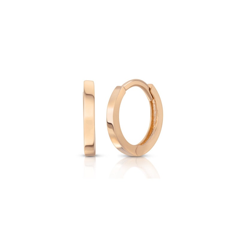 May include: Two small, gold-colored hoop earrings. One earring is open, and the other is closed. The earrings have a simple, minimalist design with a flat, rectangular profile. The earrings are displayed on a white background.