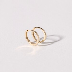 May include: Two small, gold-colored hoop earrings. The earrings are circular with a flat, rectangular profile. The earrings are displayed on a white surface, with a shadow cast behind them.