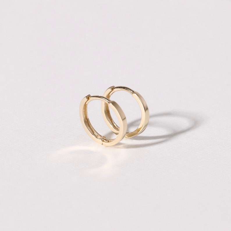 May include: Two small, gold-colored hoop earrings. The earrings are circular with a flat, rectangular profile. The earrings are displayed on a white surface, with a shadow cast behind them.
