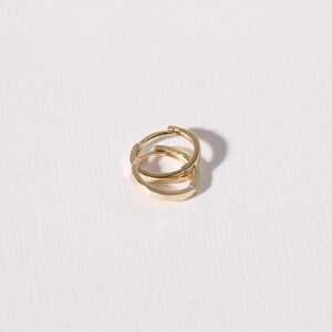 May include: Two gold-colored hoop earrings. The earrings are small, with a flat, rectangular design. The earrings are shown on a white background, with a shadow to the right.