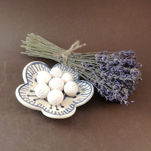 Op de afbeelding: Een wit keramisch schaaltje met een blauw bloemenpatroon, gevuld met witte keramische balletjes. Naast het schaaltje op een bruin oppervlak ligt een bos gedroogde lavendel.
