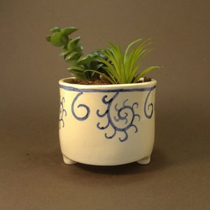 May include: White ceramic planter with blue swirl designs and three legs. The planter is filled with green succulents.