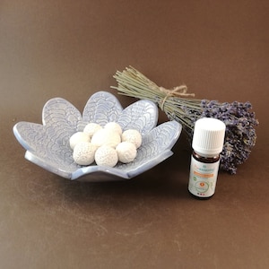 Könnte beinhalten: Eine blaue, blumenförmige Schale aus Keramik mit weißen Diffusor-Kugeln und einer kleinen Flasche Lavendel-Ätherisches Öl. Die Flasche hat einen weißen Verschluss und ein grünes Etikett mit dem Text "Pure Essential Oil Lavender"