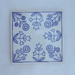May include: A white ceramic tile with a blue floral pattern. The tile features a repeating design of stylised flowers and leaves.