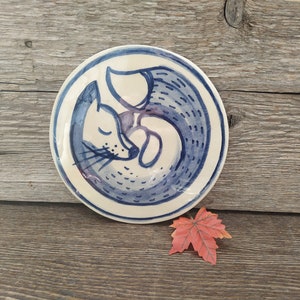 Puede incluir: Un plato de cerámica blanco con una ilustración azul y blanca de un zorro acurrucado durmiendo. El zorro está pintado en un estilo acuarela con detalles azules y blancos.