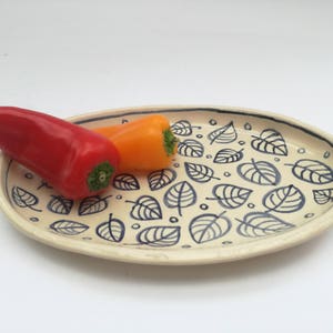 Könnte beinhalten: Eine weiße ovale Keramik-Servierplatte mit einem blauen Blatt-Muster. Zwei Paprika, einer rot und einer orange, liegen auf der Platte.