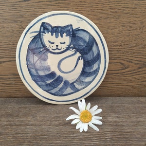 Könnte beinhalten: Ein weißer Keramikteller mit einem handgemalten Design in Blau und Weiß, das ein schlafendes, zusammengerolltes Kätzchen zeigt. Die Katze hat einen blau-weiß gestreiften Schwanz und einen weißen Bauch.