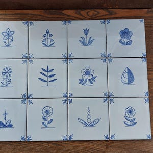 Puede incluir: Un conjunto de doce baldosas de cerámica blancas dispuestas en una cuadrícula. Cada baldosa presenta un diseño floral azul dibujado a mano. Las baldosas son probablemente para uso decorativo.