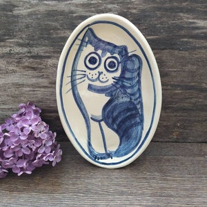 Op de afbeelding: Een wit ovale keramische schaal met een handgeschilderd ontwerp in blauw en wit van een kat. De kat heeft grote ogen en een vriendelijke uitdrukking.