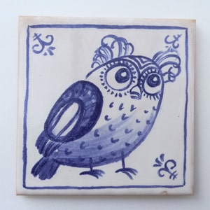 May include: A blue and white ceramic tile with a hand-painted design of an owl. The owl has large eyes and a detailed pattern of dots and lines. The tile is framed with a simple blue border.