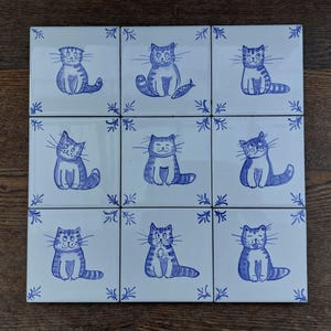 Puede incluir: Nueve azulejos de cerámica blanca con ilustraciones de gatos azules y blancos. Cada azulejo presenta un gato diferente con una expresión y una pose únicas. Los azulejos están dispuestos en una cuadrícula de 3x3.