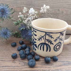 May include: A cream ceramic mug with a dark blue floral pattern and a handle. Blueberries and blue thistle flowers are arranged around the mug on a wooden surface. The mug is approximately 10 cm tall.