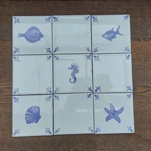 Könnte beinhalten: Neun quadratische Keramikfliesen mit hellblauem Hintergrund und blauen Illustrationen von Meereskreaturen. Die Fliesen zeigen einen Fisch, ein Seepferdchen, einen Seestern und eine Muschel. Jede Fliese hat ein blaues Blumeneckdesign.