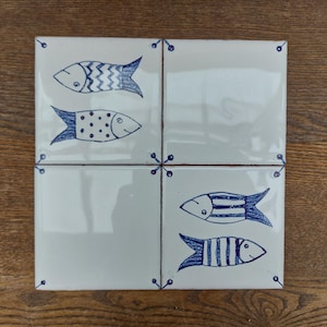 Puede incluir: Cuatro baldosas cuadradas blancas, cada una con un diseño de pez azul dibujado a mano. Los peces tienen diferentes patrones, incluyendo zigzags, puntos y rayas. Acentos decorativos azules en las esquinas e intersecciones.