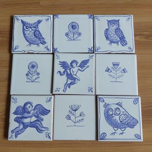 Peut inclure: Neuf carreaux de céramique carrés avec des motifs bleus et blancs. Les carreaux présentent des illustrations de hiboux, d'anges et de motifs floraux. Les carreaux sont disposés en grille de trois par trois sur une surface en bois.
