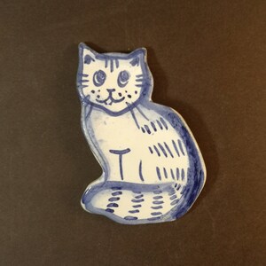 Könnte beinhalten: Handbemalte Keramik-Katzenfigur mit blau-weißer Glasur. Die Katze sitzt, mit großen Augen und einem lächelnden Gesicht. Der Körper hat gestreifte Details. Die Figur ist etwa 10 cm hoch.